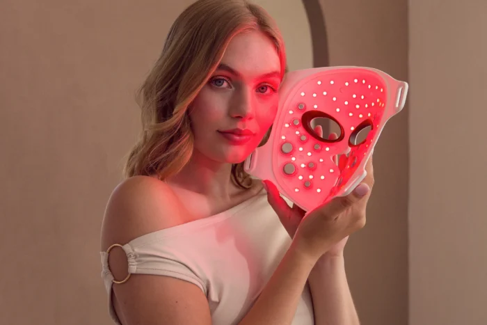 Uśmiechnięta kobieta siedząca na kanapie i trzymająca maskę LED z EMS do domowego liftingu twarzy i likwidacji chomików